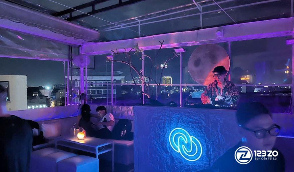 Top 4 Quán Rooftop Nổi Tiếng Tại Quận Phú Nhuận View Đỉnh Cho Tín Đồ Sống Ảo 8 Quán rooftop tại Quận Phú Nhuận với ánh sáng tím, khu vực ghế sofa và một DJ đang chơi nhạc ở quầy, có tầm nhìn ra thành phố đêm.